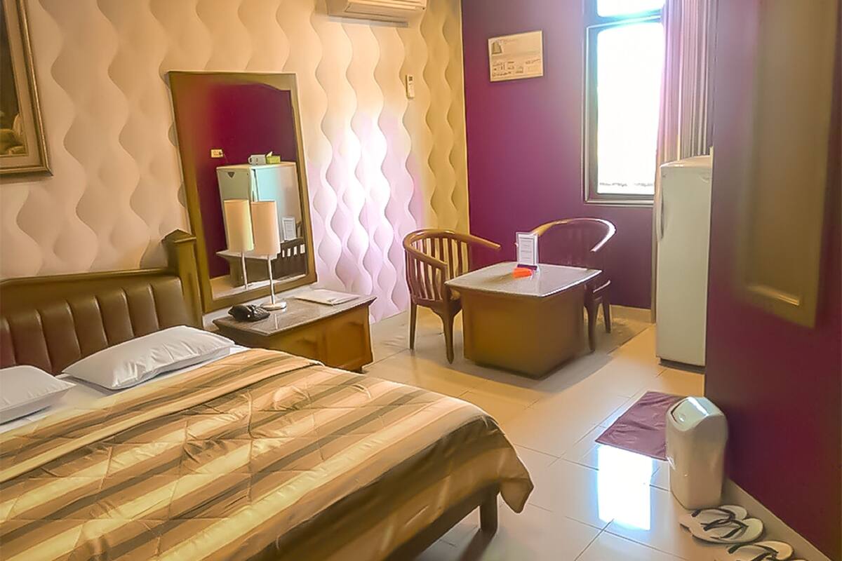 A hotel room with a bed, a desk, a chair, a mirror, and a refrigerator.