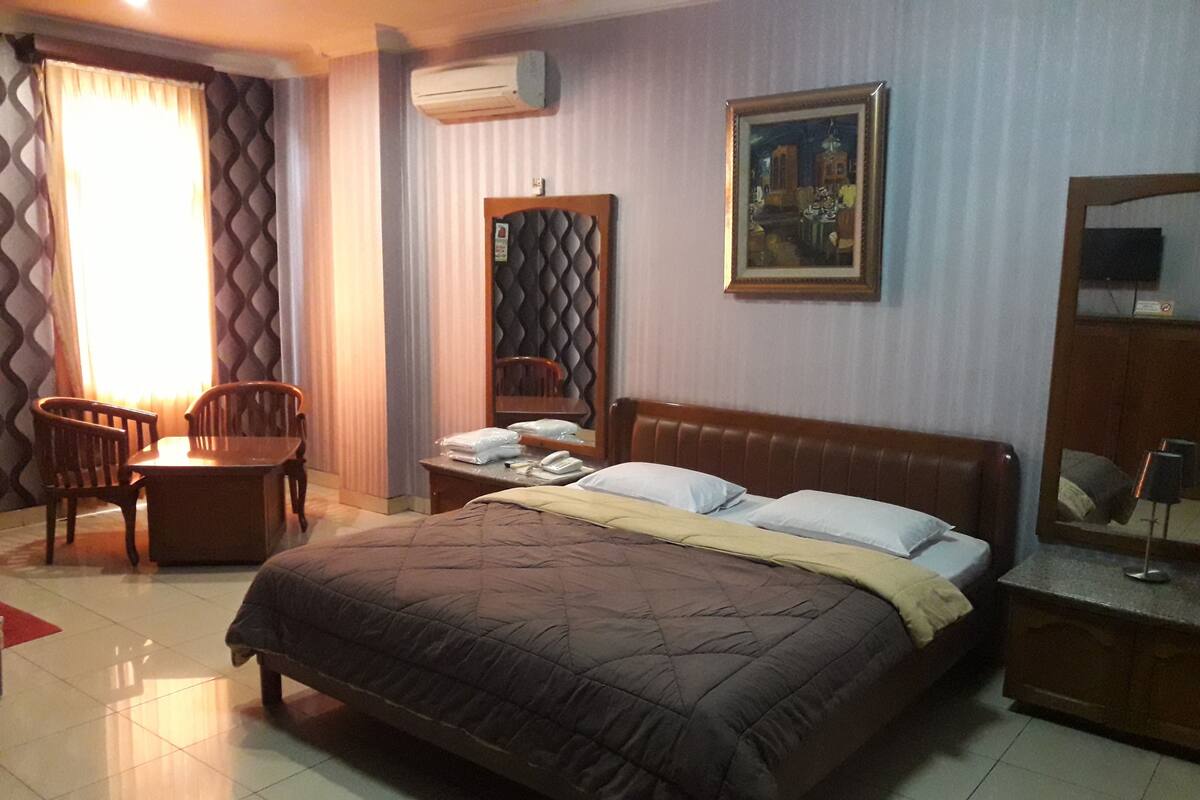 A hotel room with a bed, a desk, a chair, an air conditioning unit, a painting, and a mirror.