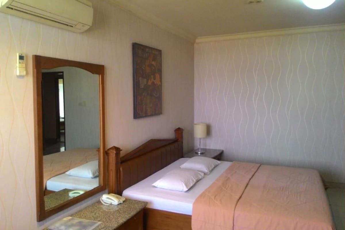 Une chambre d’hôtel comprenant un lit, une table de chevet, un miroir, un climatiseur fixé au mur et un papier peint à motifs.