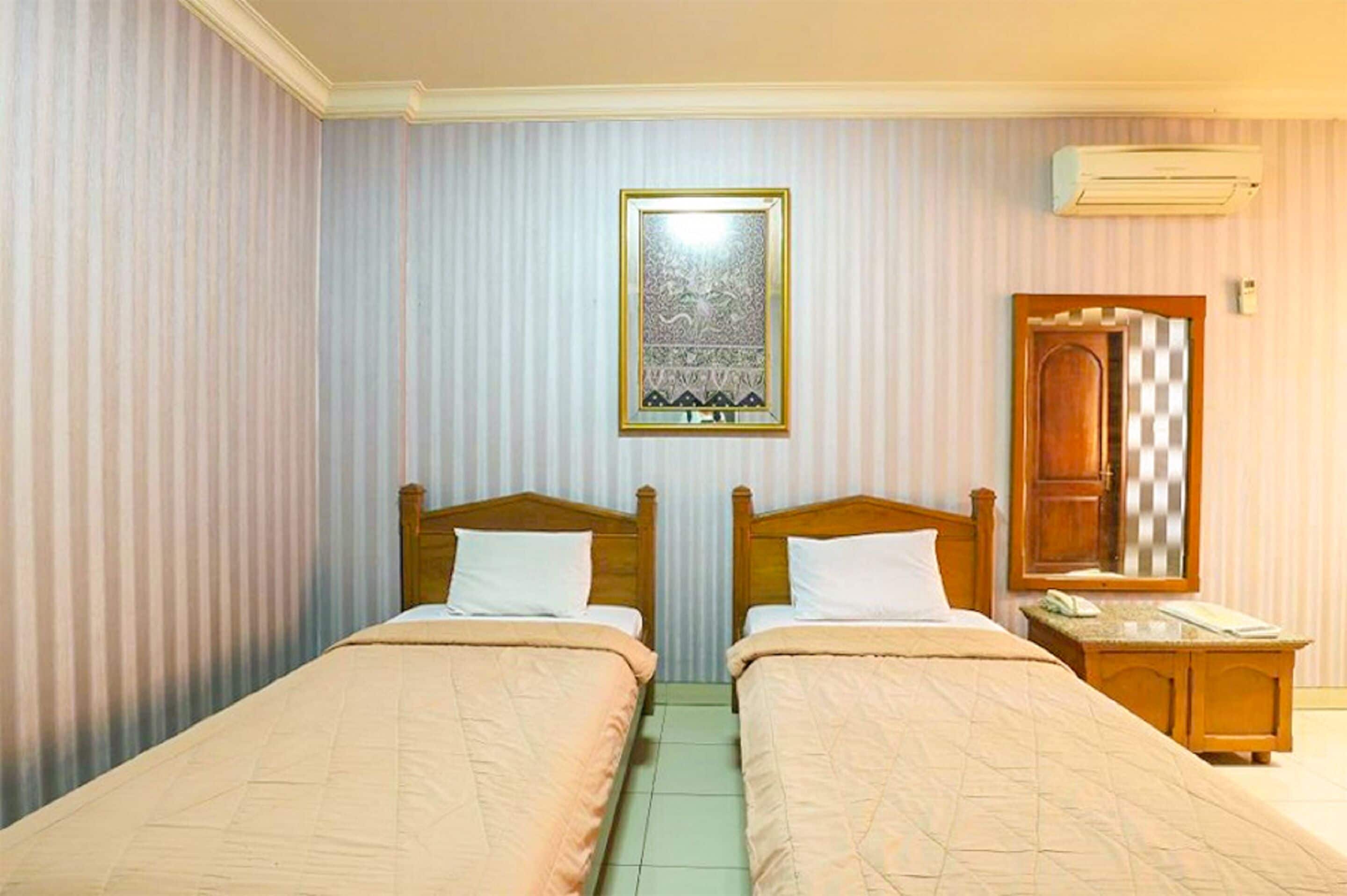 A hotel room with two single beds, a wooden nightstand with a telephone, a wall-mounted air conditioner, and a framed artwork above the beds.