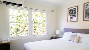 A bedroom with a large window, a bed, a bedside table, and two framed pictures on the wall. - Lotus Stay Manly - Apartment 31F (Manly)