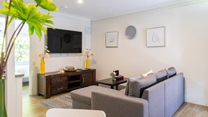 A modern living room with a grey sofa, a small table, a TV mounted on the wall, and a decorative vase with flowers. - Lotus Stay Manly - Apartment 31F (Manly)