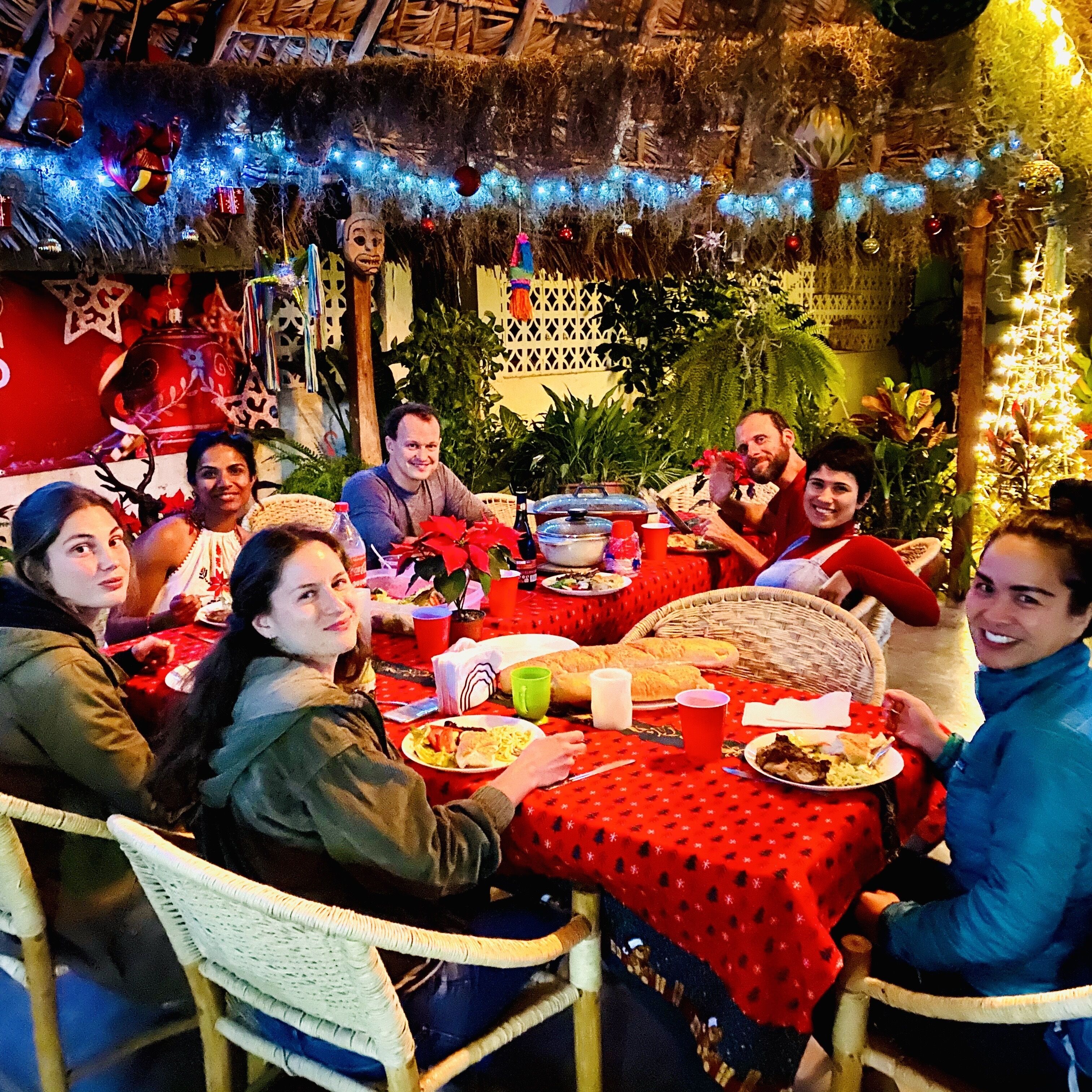A group of people dining at a table with a red tablecloth, surrounded by festive decorations and plants.