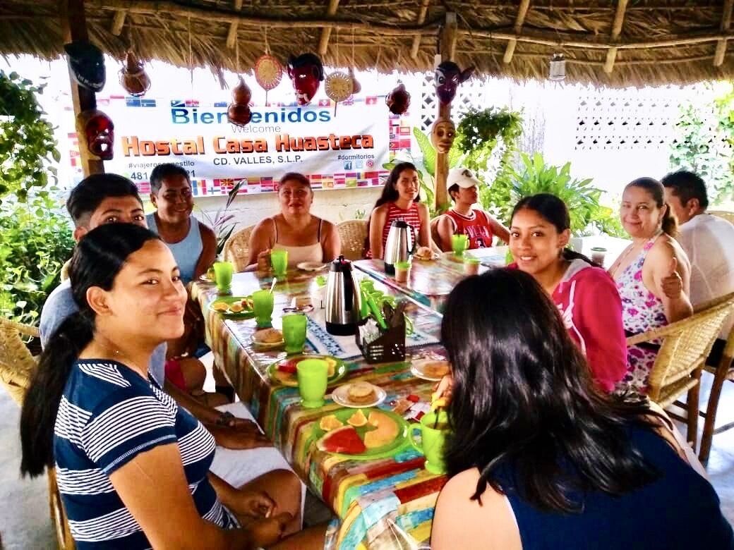 A group of people seated around a table with a colorful tablecloth, under a thatched roof structure.