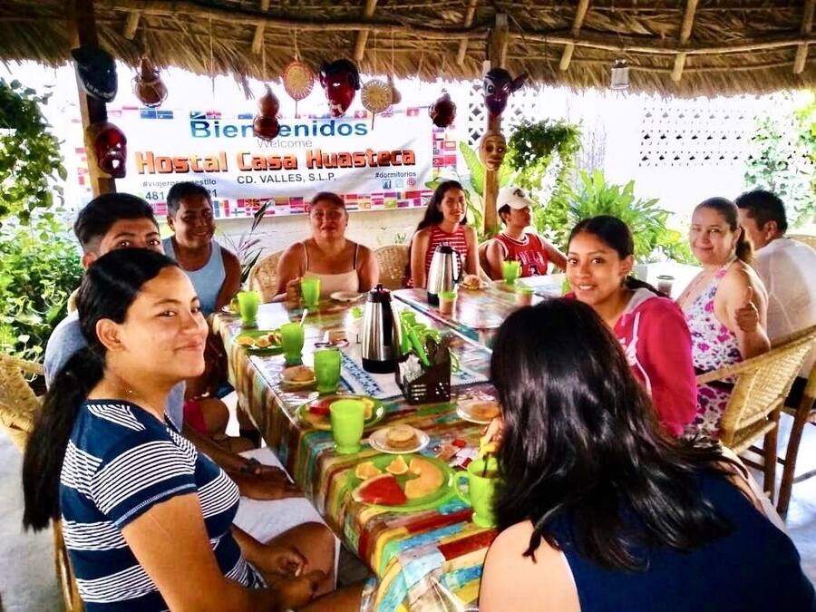 A group of people seated around a table with a colorful tablecloth, under a thatched roof structure.