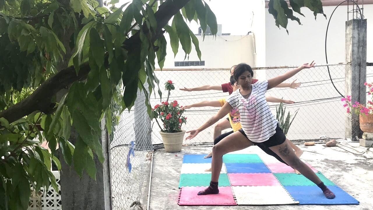 Eine Person führt eine Yoga-Pose auf einer Matte im Freien durch, im Hintergrund ist ein Zaun und Kübelpflanzen zu sehen.