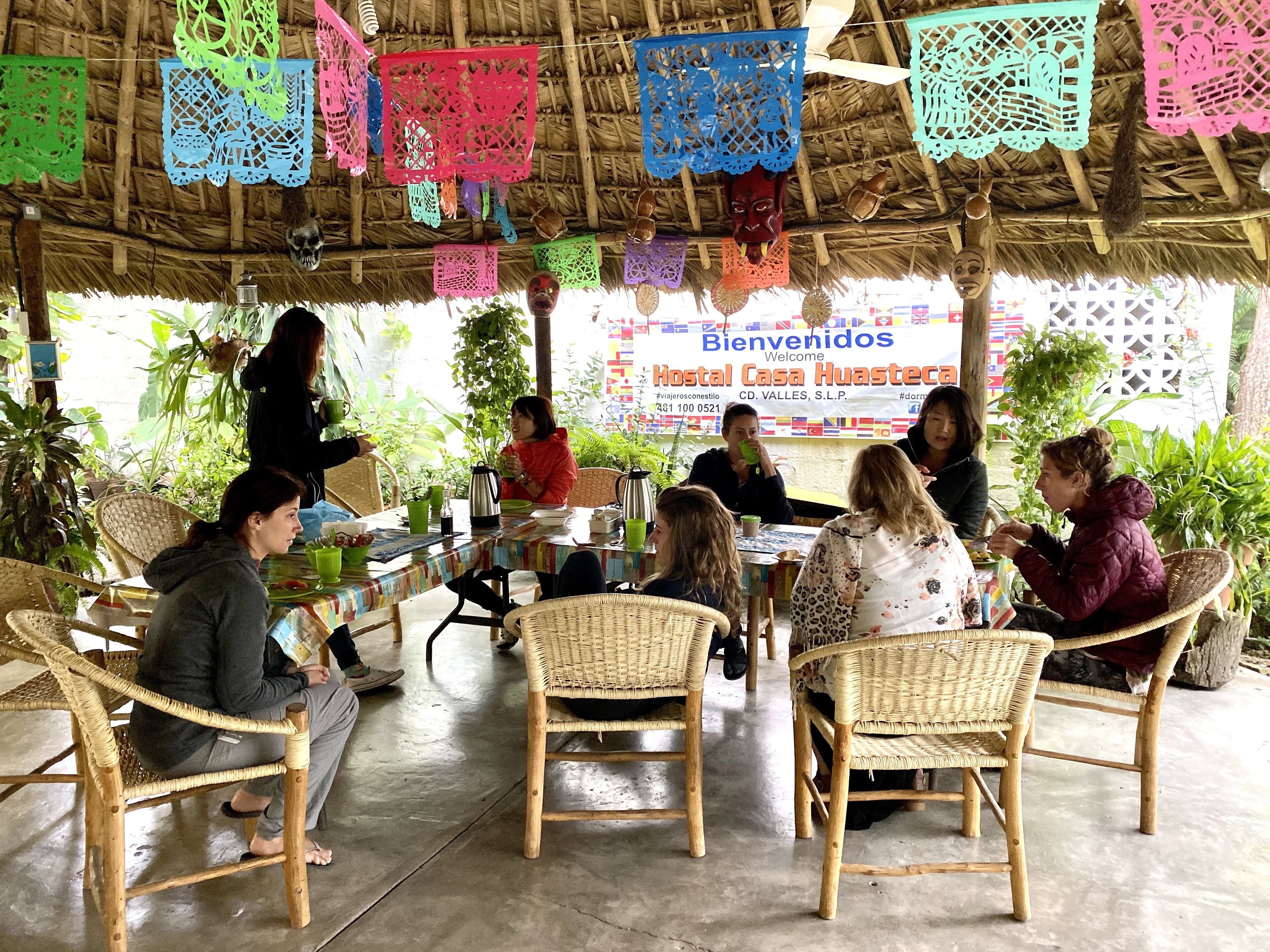 A group of people seated at a table under a thatched roof, with colorful paper decorations hanging above.