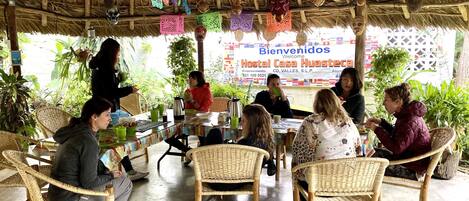 A group of people seated at a table under a thatched roof, with colorful paper decorations hanging above.