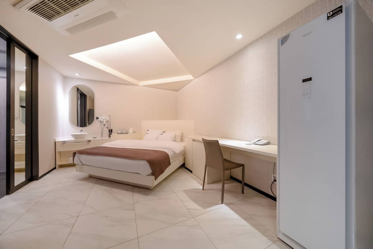 A modern hotel room with a bed, desk, chair, and a refrigerator.