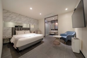 Una habitación de hotel moderna con una cama grande, una mesita de noche, una pequeña mesa redonda, un sillón azul, un espejo y un equipo de aire acondicionado instalado.
