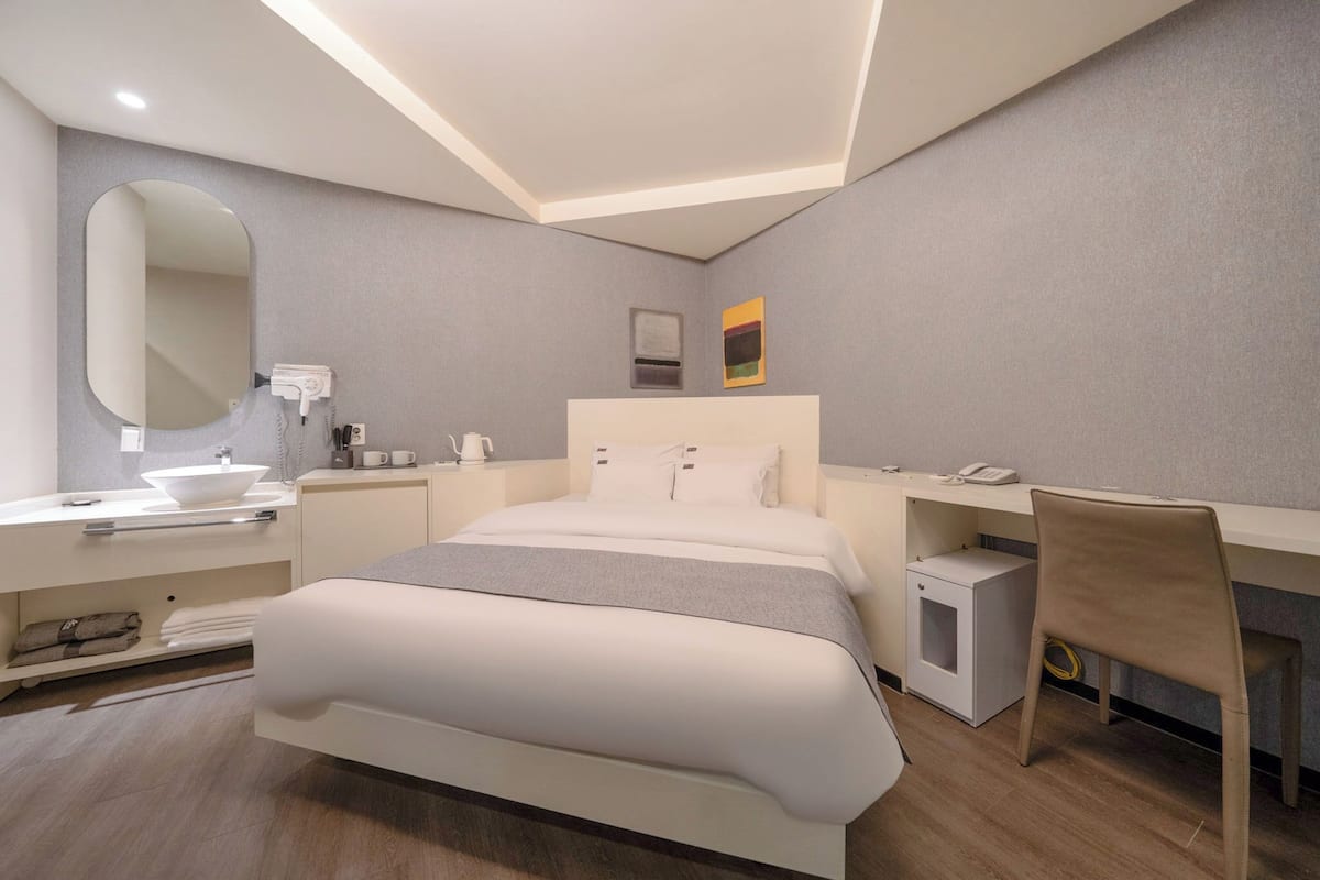 A modern hotel room with a large bed, a bedside table, a desk with a chair, a mirror, and a bathroom area with a sink and towel rack.