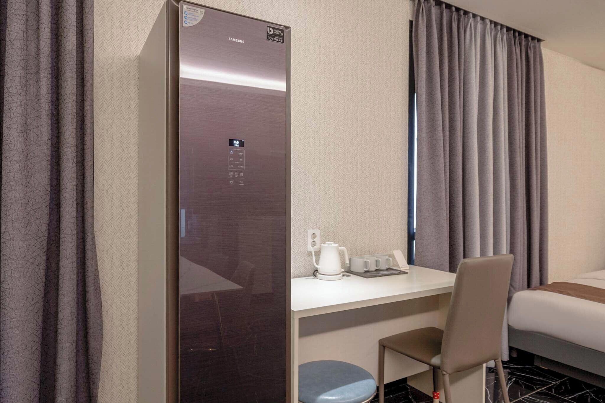 Quarto de hotel com cama, escrivaninha com cadeira, pequena mesa com chaleira e chávenas, e frigorífico fixado na parede.
