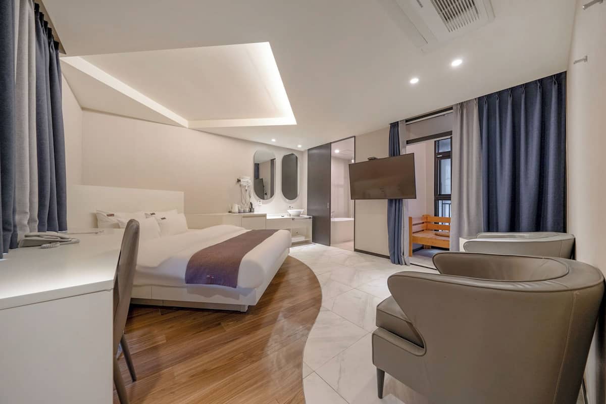 A modern hotel room with a bed, a desk, a chair, a TV, and a bathroom visible through an open door.