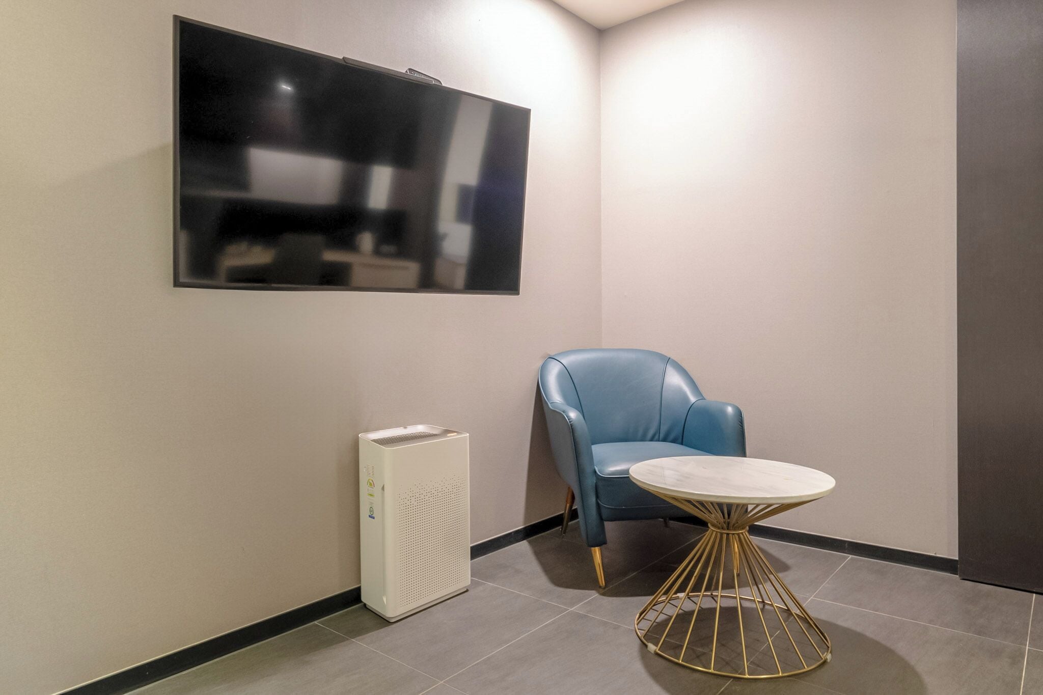 A corner room with a blue chair, a small round table with a gold base, and a large flat-screen TV mounted on the wall.