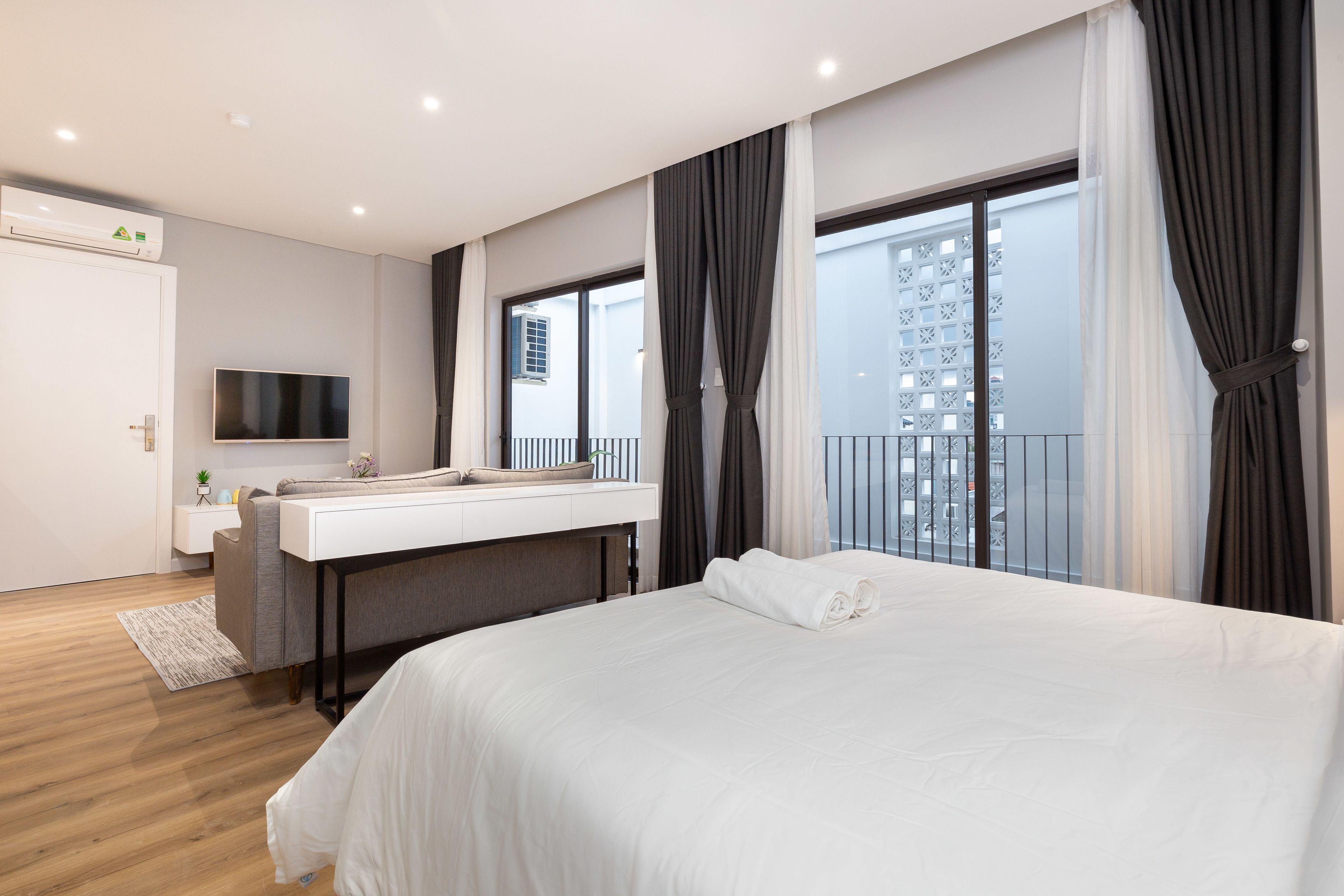 A modern hotel room with a large bed, a desk with a flat-screen TV, a balcony with curtains, and a view of the city.