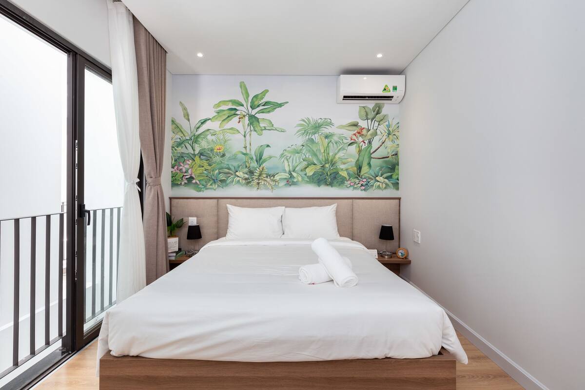 Une chambre d’hôtel avec un grand lit, une fresque représentant des plantes tropicales sur le mur, un climatiseur et un balcon avec vue.