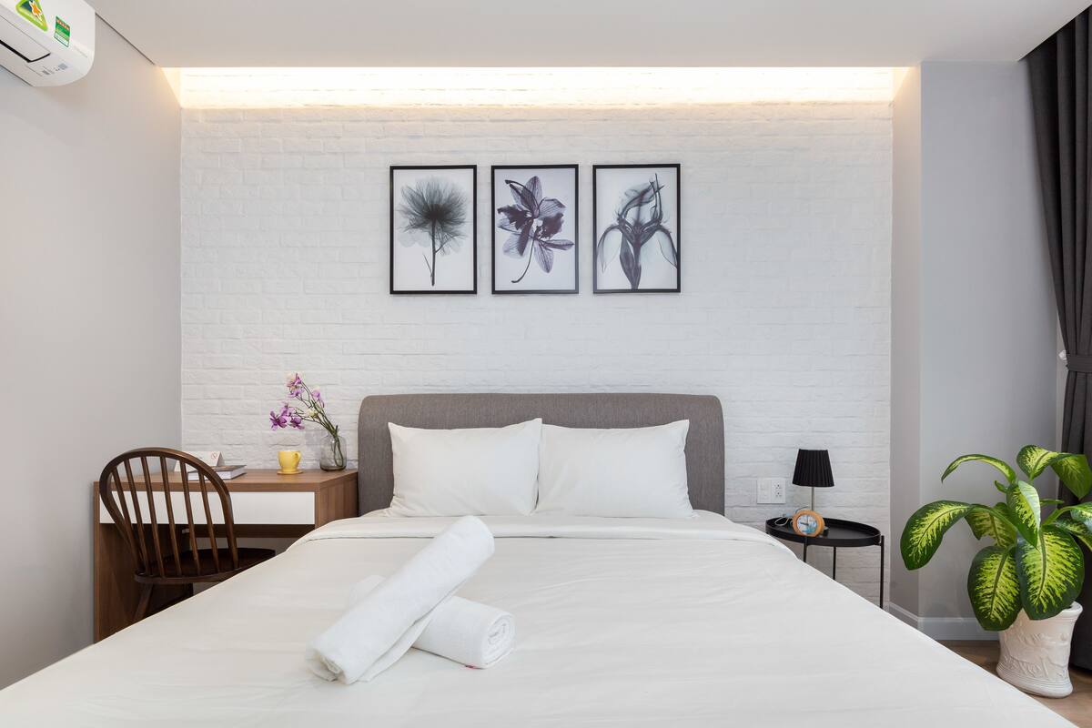 Une chambre d’hôtel bien rangée, avec un lit, un bureau, une chaise, une table de chevet, une lampe et une plante en pot.