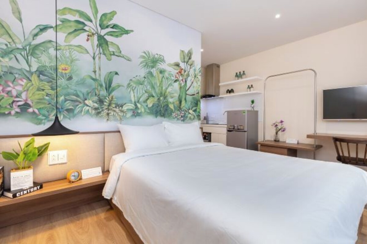 Une chambre avec un lit, une table de chevet ornée d’une plante, un téléviseur fixé au mur, et un coin cuisine à l’arrière-plan.