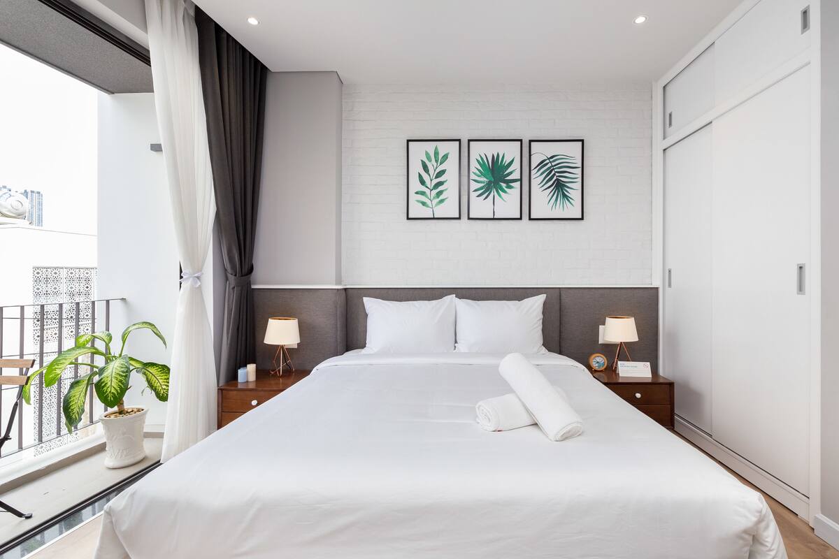Une chambre d’hôtel moderne avec un grand lit, deux tables de chevet avec des lampes, une plante en pot et des reproductions encadrées de motifs botaniques accrochées au mur.