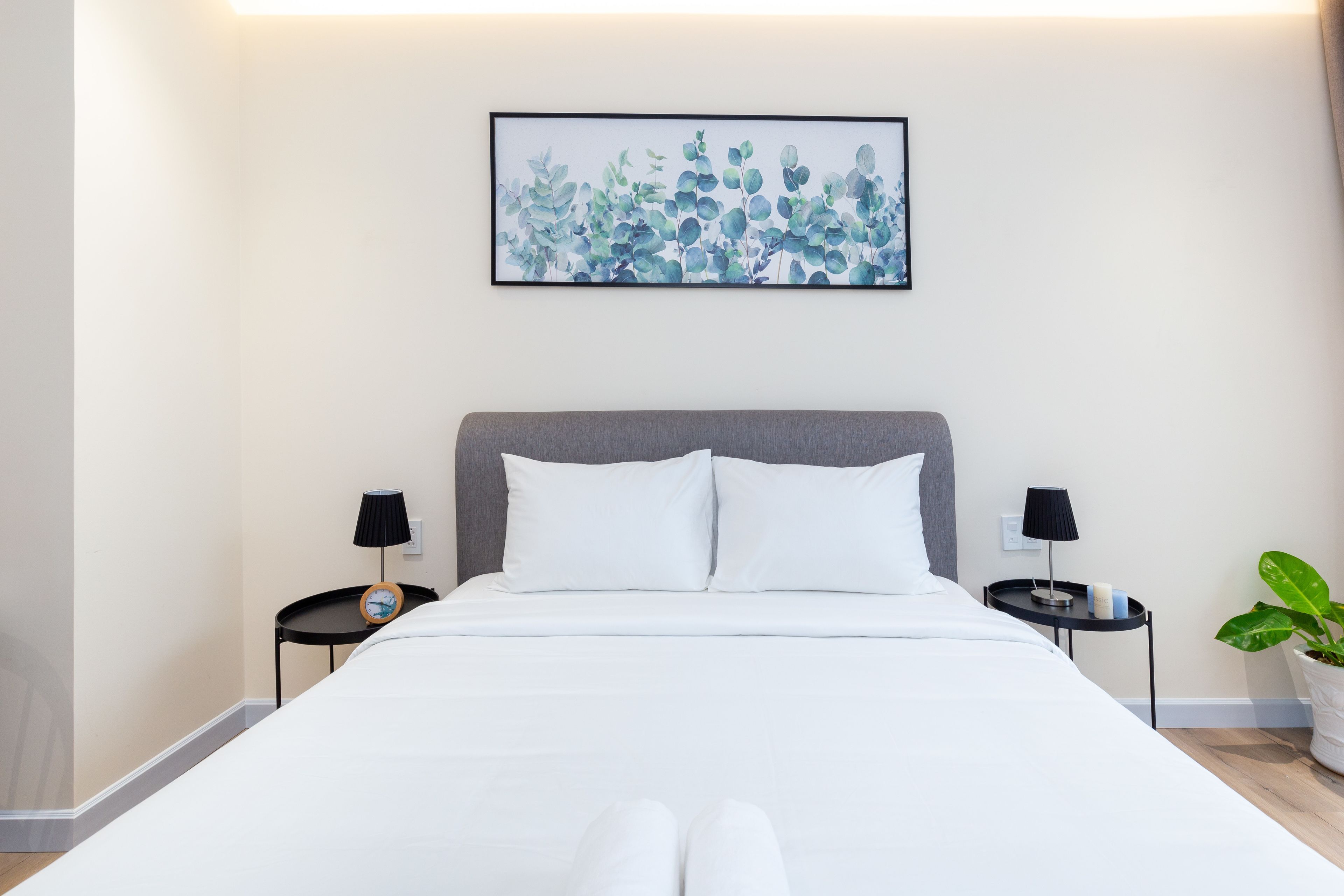 Cama bem arrumada com roupa de cama branca, cabeceira cinza e duas mesas de cabeceira com abajures. Uma obra de arte emoldurada está pendurada acima da cama, e um vaso com planta é visível no lado direito.
