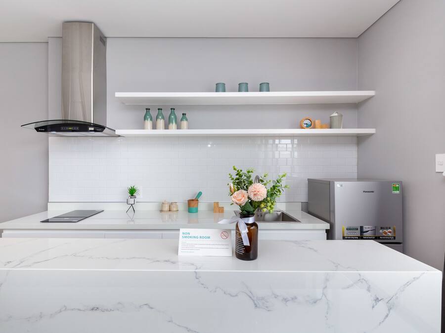 Eine moderne Küche mit weißen Arbeitsplatten, einer Edelstahl-Dunstabzugshaube und einem Kühlschrank. Darüber befinden sich Regale mit Flaschen und eine Vase mit Blumen. Ein Schild weist darauf hin, dass es sich um einen Nichtraucherraum handelt.
