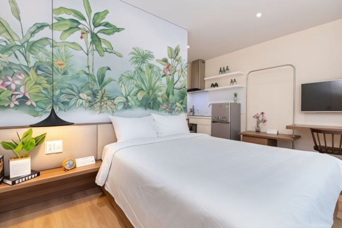 Une chambre d’hôtel comprenant un lit, un bureau, une chaise, un téléviseur, un coin cuisine et un mur orné d’une fresque représentant des plantes tropicales.