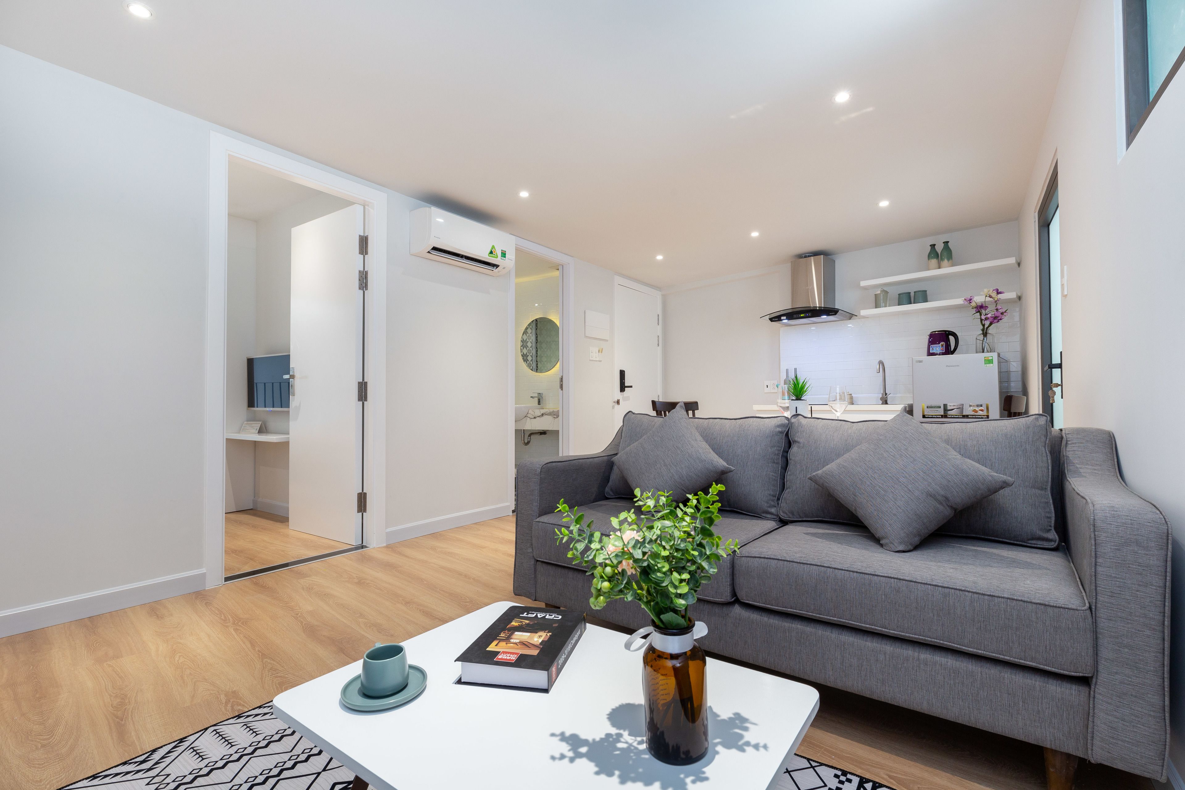 A modern living room with a grey sofa, a coffee table with a book and a vase, and a kitchen in the background.