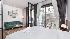Una habitación de hotel moderna con una cama grande, un sofá verde, una mesita y un balcón con mesa y sillas.