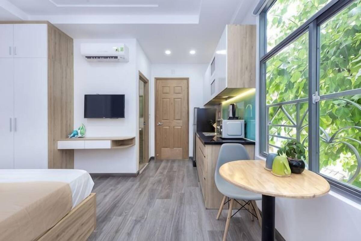 A modern hotel room with a bed, a TV, a small kitchen area, and a large window with a view of trees.