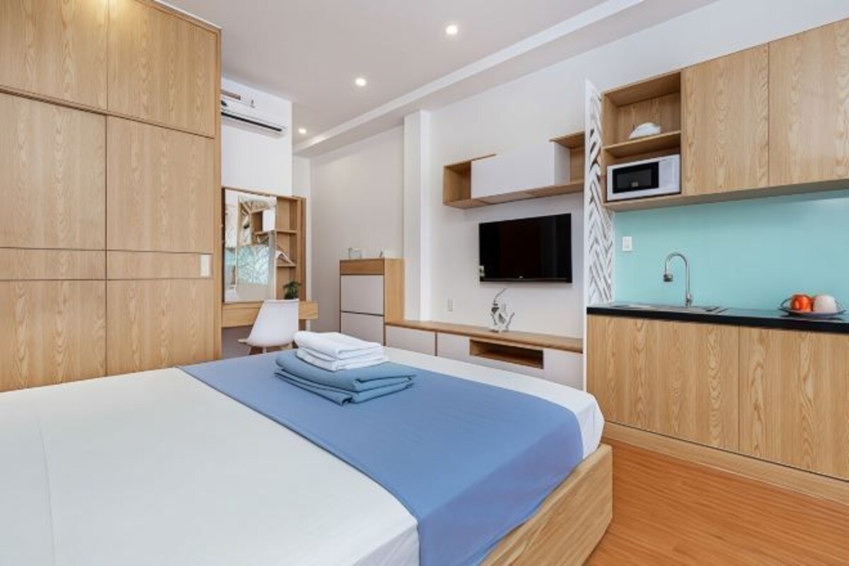 A modern hotel room with a bed, a TV, a kitchenette, and wooden cabinetry.