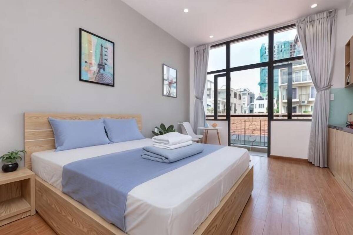 A modern hotel room with a large bed, a small bedside table, a nightstand, a window with a view of buildings, and a balcony.