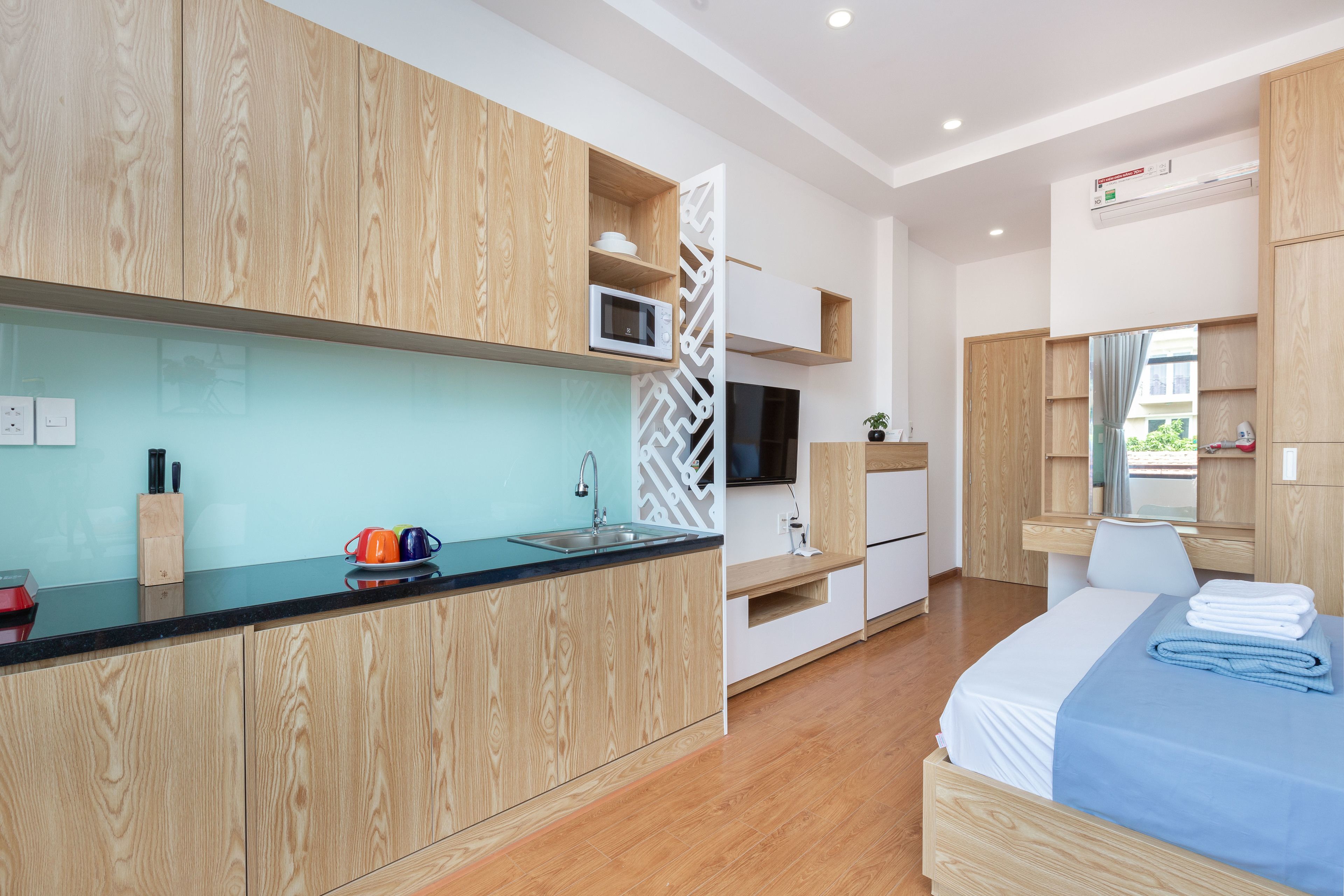 A modern hotel room with a wooden floor, a bed with a blue blanket, a small kitchenette with a microwave, and a TV.