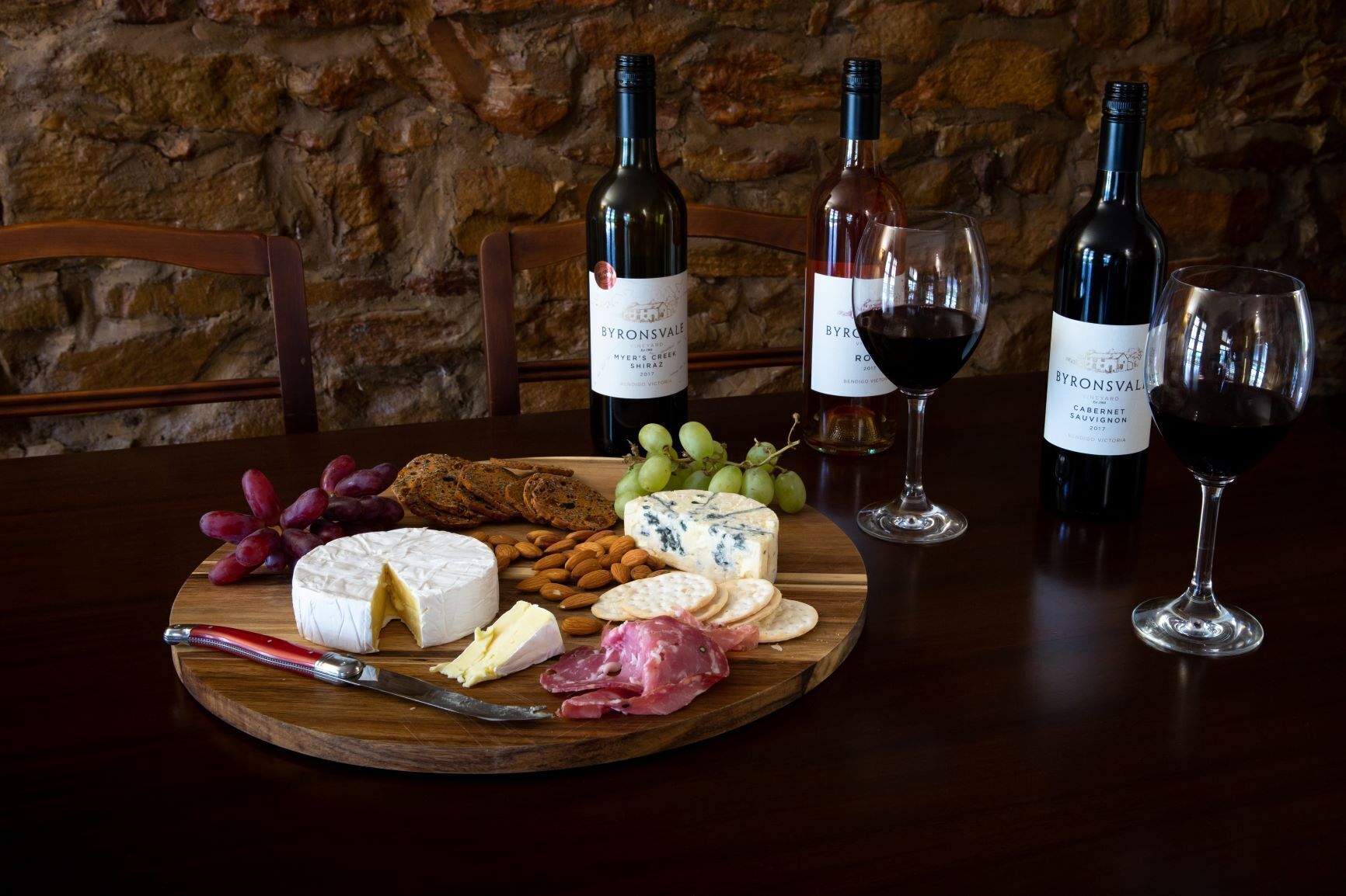 A wooden board with cheese, crackers, and grapes, accompanied by wine bottles and glasses on a table.