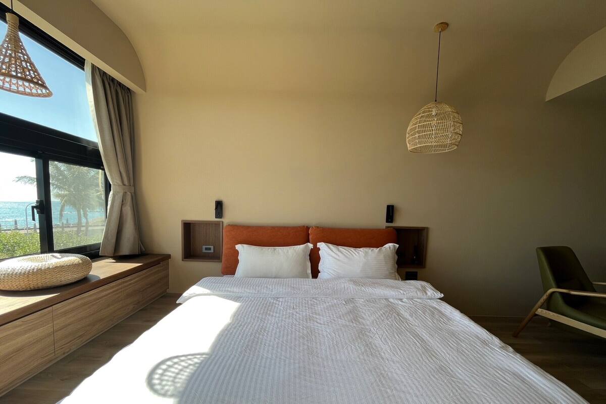 A bedroom with a large bed, a window with a view of the sea, a bedside table, and a hanging light fixture.