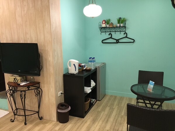 Una habitación con televisión, tetera, una pequeña mesa con cubierta de vidrio, una silla, un cubo de basura, un estante con plantas y una lámpara colgante.