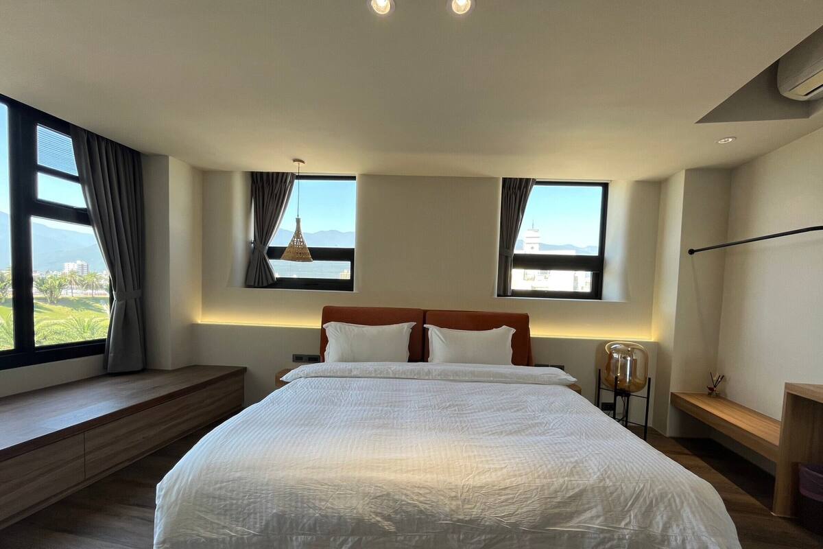 A modern bedroom with a large bed, two windows with curtains, a bedside table, and a view of the outdoors.
