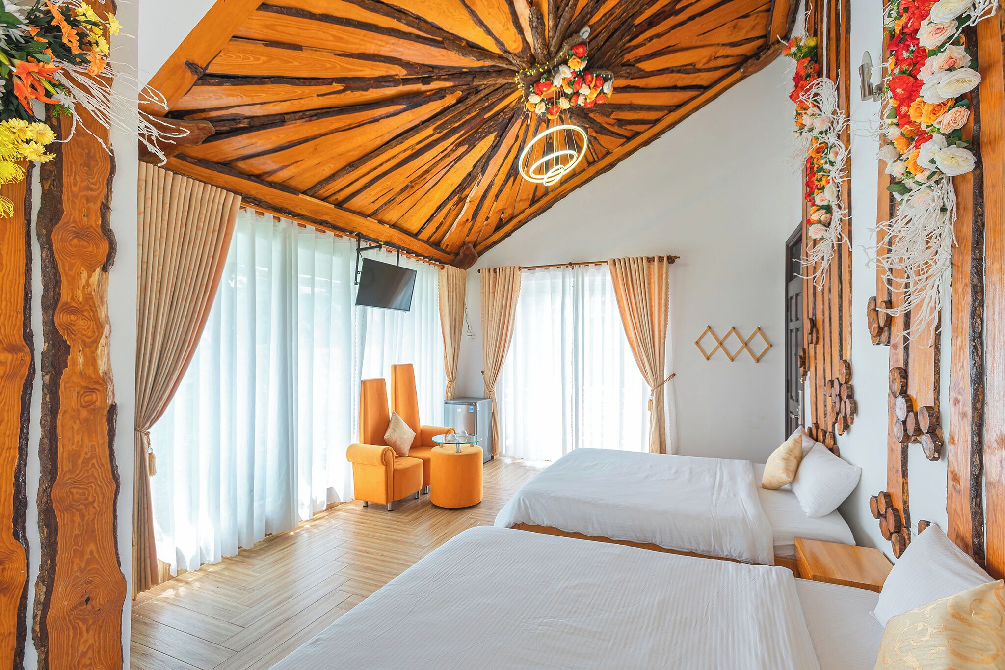 A hotel room with two beds, a wooden ceiling, curtains, a TV, and a floral decoration.