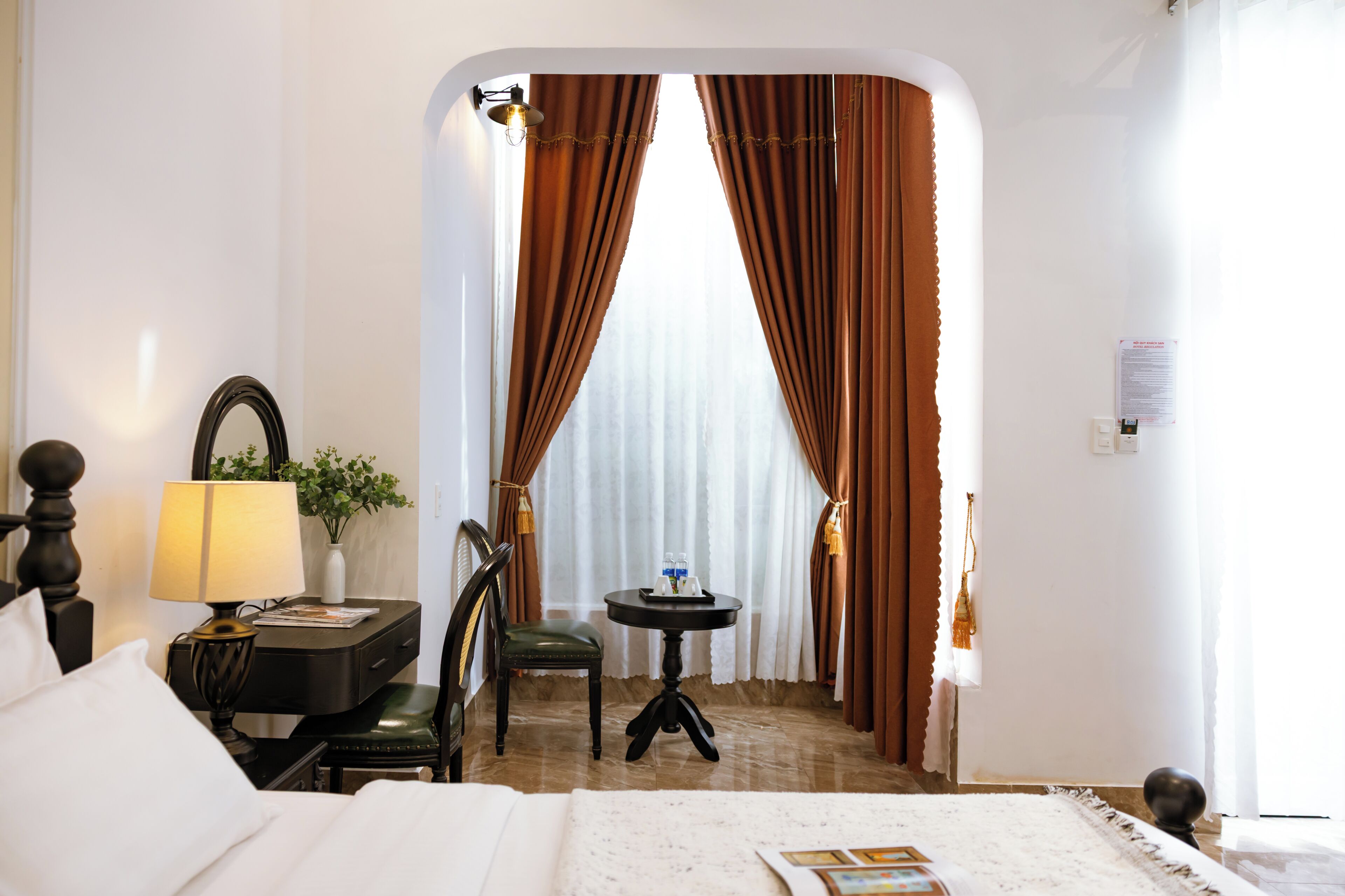 A hotel room with a bed, a desk with a lamp, a chair, a small table with a plant, and a view through curtains.