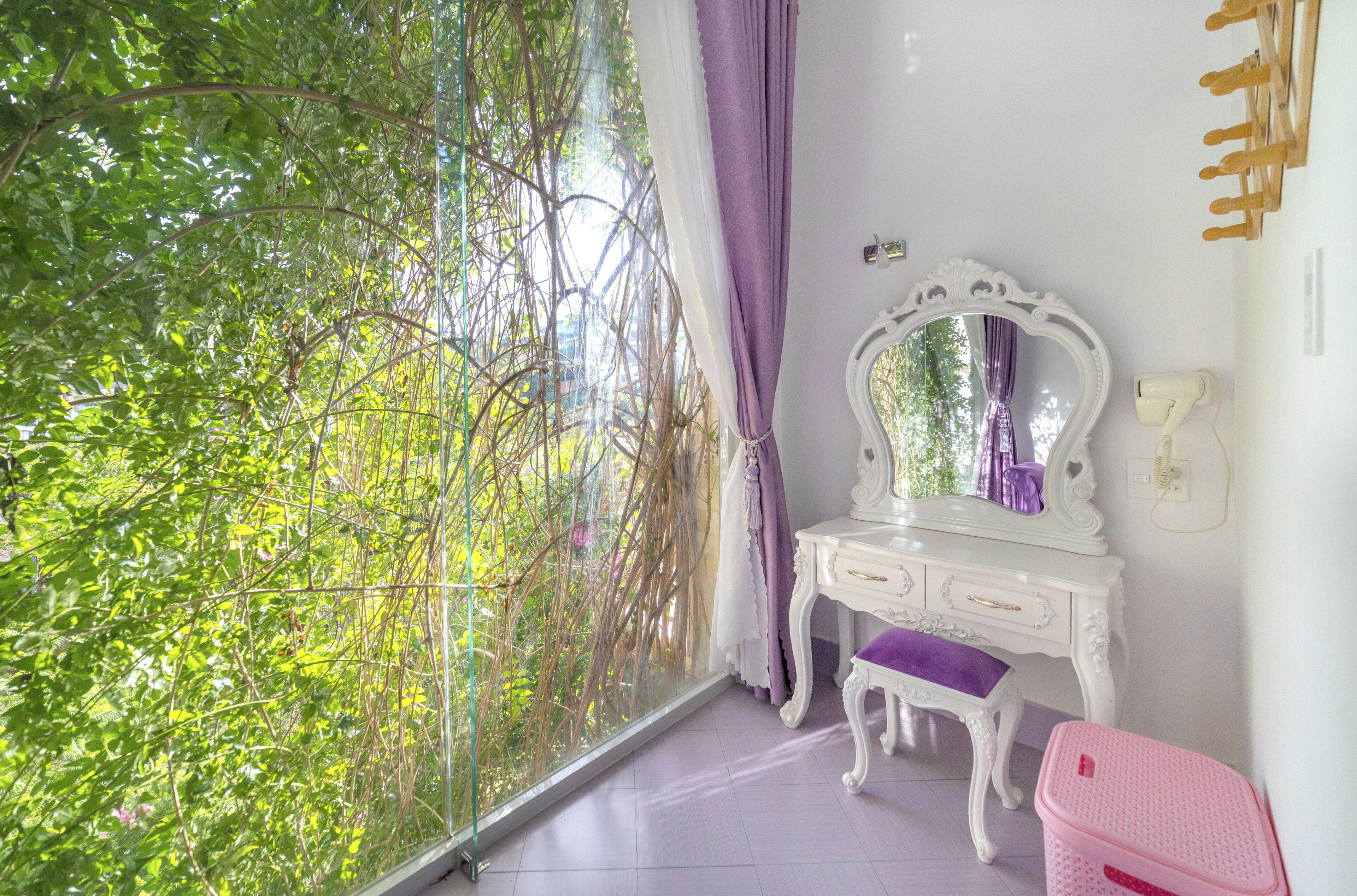 Ein Zimmer mit einem großen Fenster, einem weißen Schminktisch mit Spiegel, einem pinkfarbenen Koffer und einem weiß montierten Föhn.
