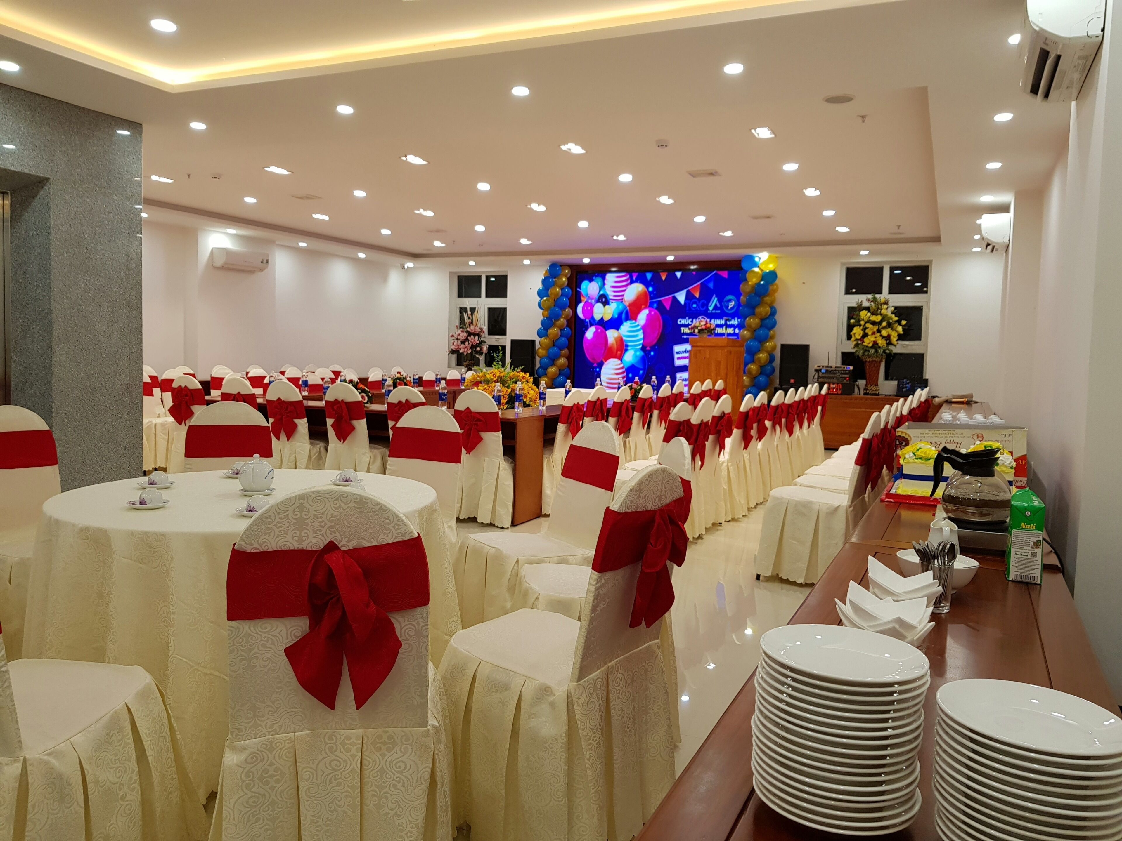 A banquet hall with round tables, white chairs with red sashes, and a stage with a colorful backdrop.