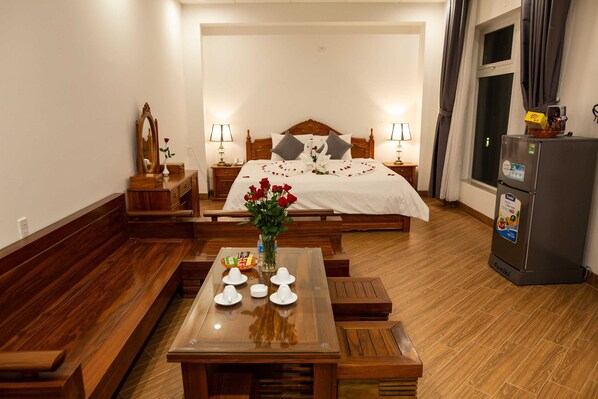 Quarto de hotel com um banco de madeira, uma mesa de jantar com pratos e um vaso de flores, uma cama com almofadas, uma mesa de cabeceira, uma luminária e um refrigerador.