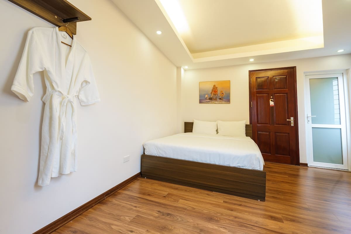 Un dormitorio con piso de madera, una cama con ropa de cama blanca, una puerta de madera y un cuadro en la pared.