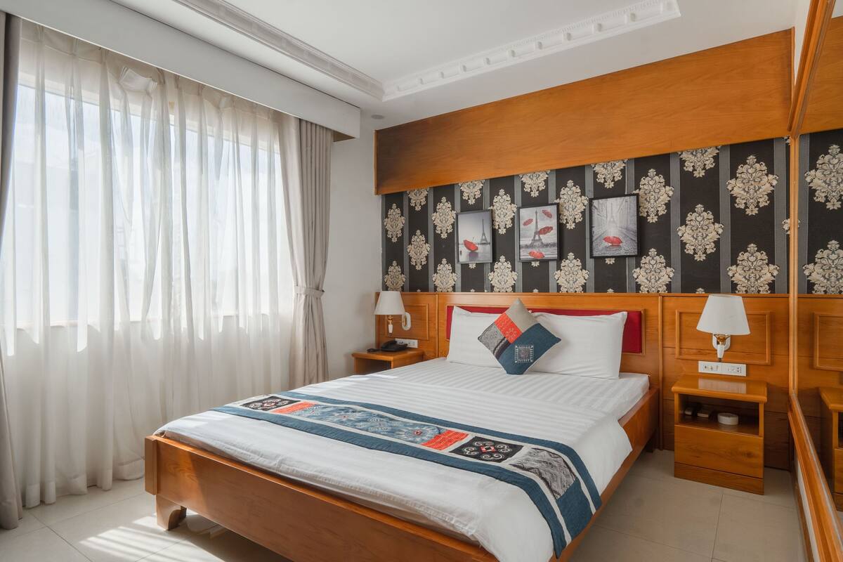 Hotelkamer met een houten bed, een nachtkastje met lamp, een wand met ingekaderde foto's en dunne gordijnen.