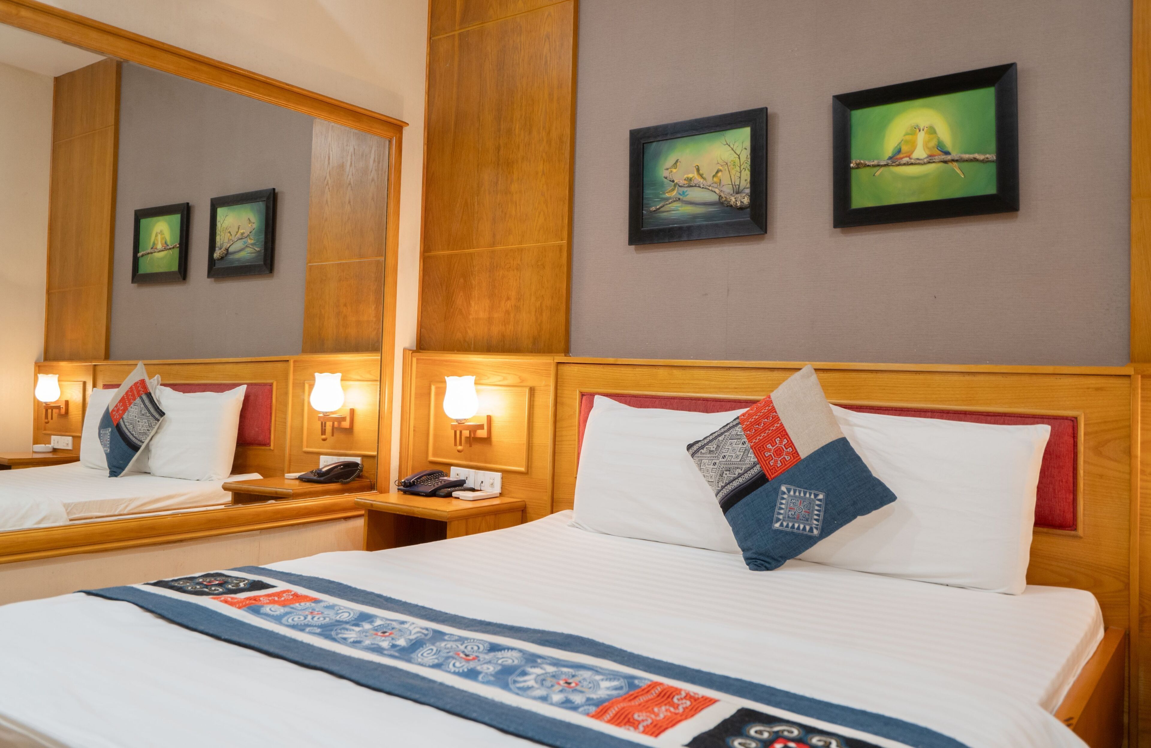 Une chambre d’hôtel avec deux lits, des têtes de lit en bois et des tables de chevet avec des lampes. Des tableaux encadrés ornent le mur et un coussin décoratif est posé sur le lit.