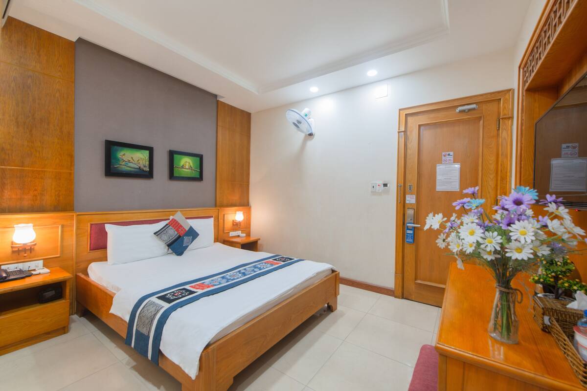Een hotelkamer met een houten bed, bedlampen, een nachtkastje, een bureau met een bloevas en twee ingelijste foto's aan de muur.