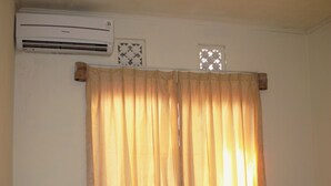 A room with a bed, a chair, a small table, an air conditioner, and a window with a curtain. - Bale Gerupuk (Kuta)