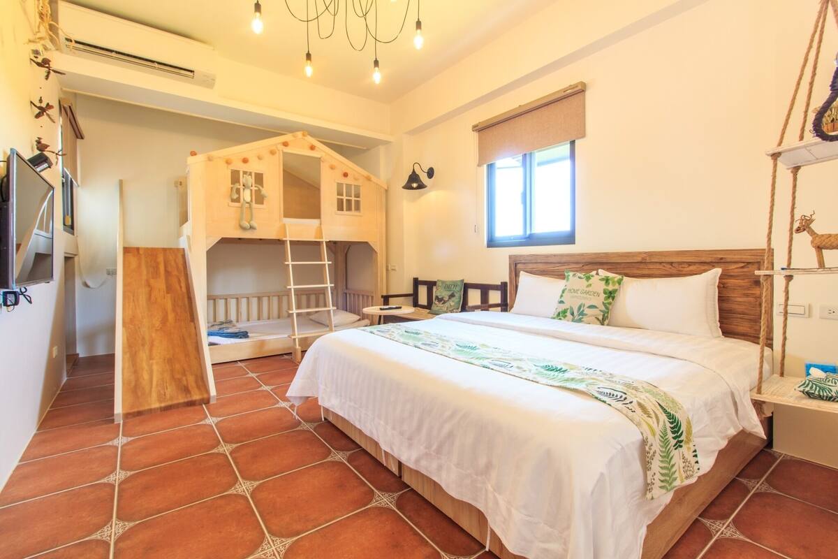 A bedroom with a bunk bed, a wooden playhouse, a ladder, a bedside table with a lamp, and a window with a view.