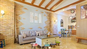 A living room with a sofa, coffee table, and children's stools. There is a brick wall with a partial brickwork design, a wooden ceiling, and a dining area with chairs and a table. - BJ Classic Town (Wujie)