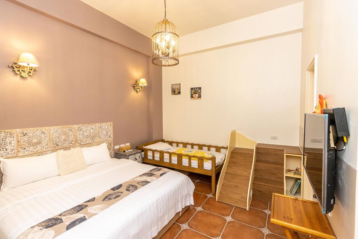 A bedroom with a large bed, a wooden bench, a TV mounted on the wall, and a chandelier.