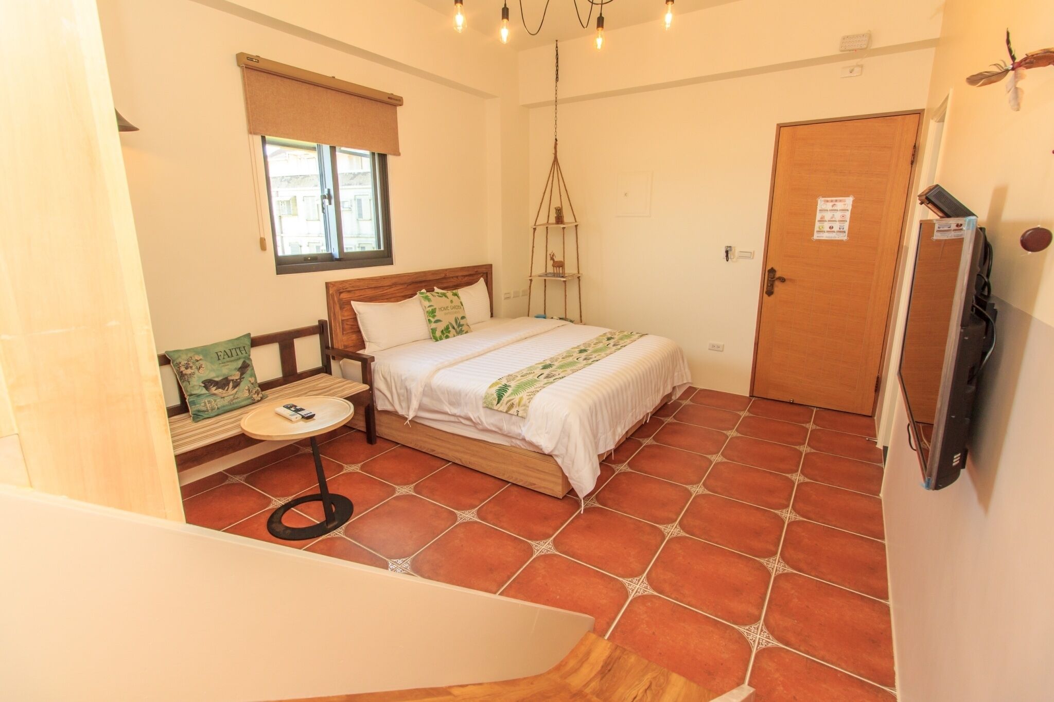 A bedroom with a bed, a small table, a chair, a window with a blind, a door, and a wall-mounted shelf.